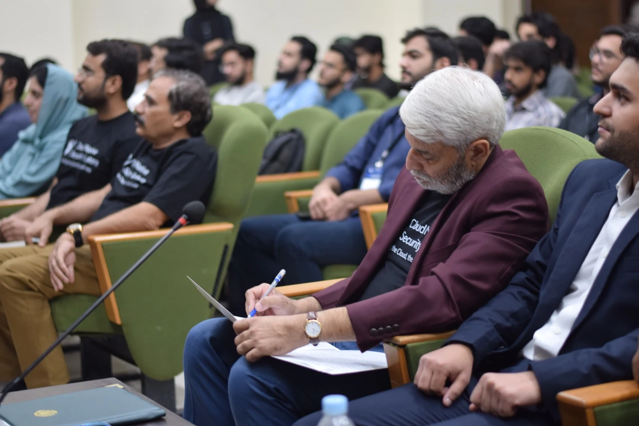 A front-row guest in a burgundy blazer writes notes during the session.