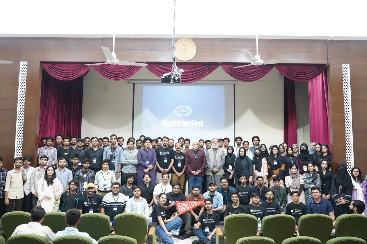 A large group of Hacktoberfest participants pose beneath the event screen at Lahore Garrison University.