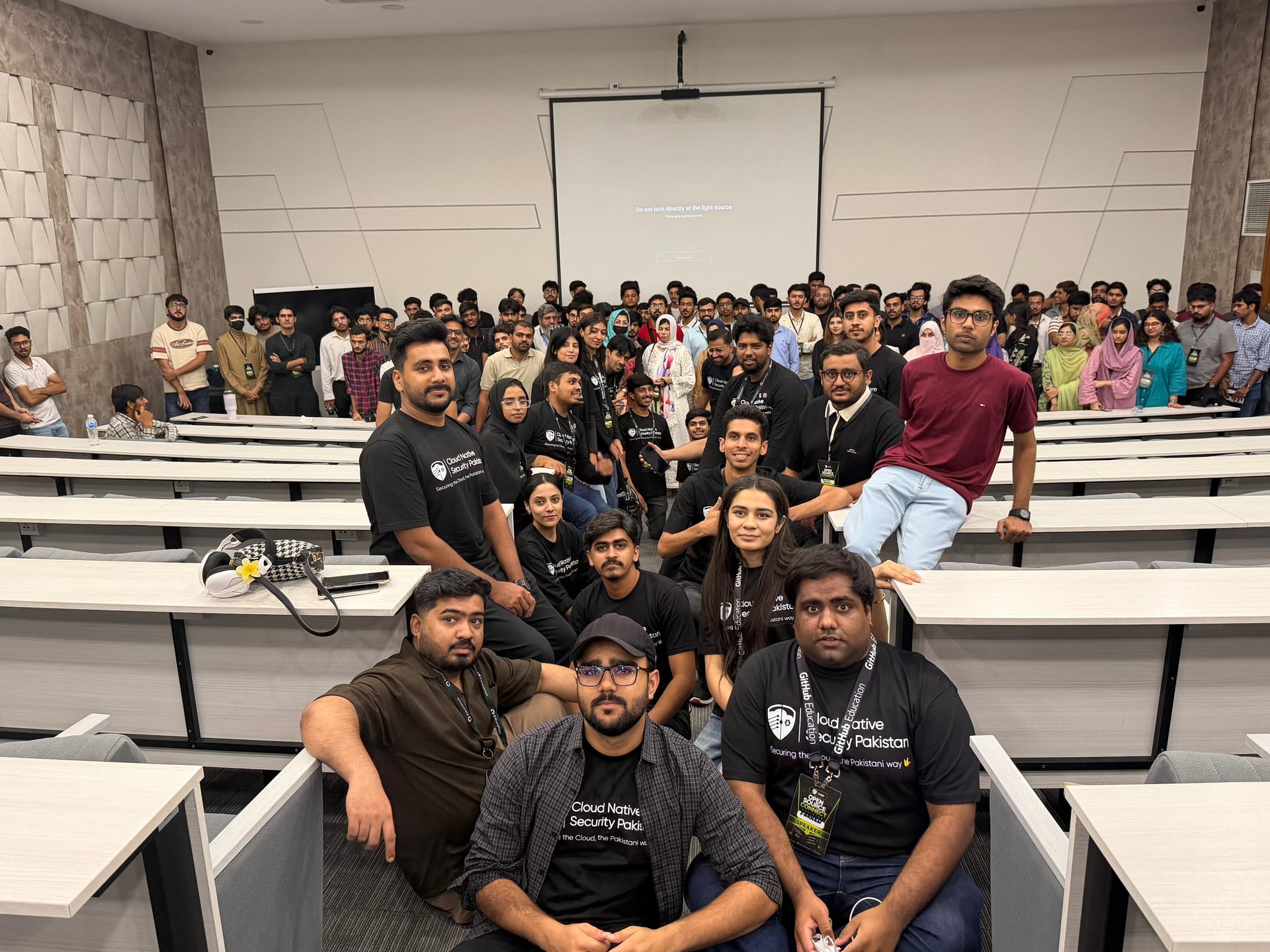 A large group of attendees and organizers poses between the lecture hall desks